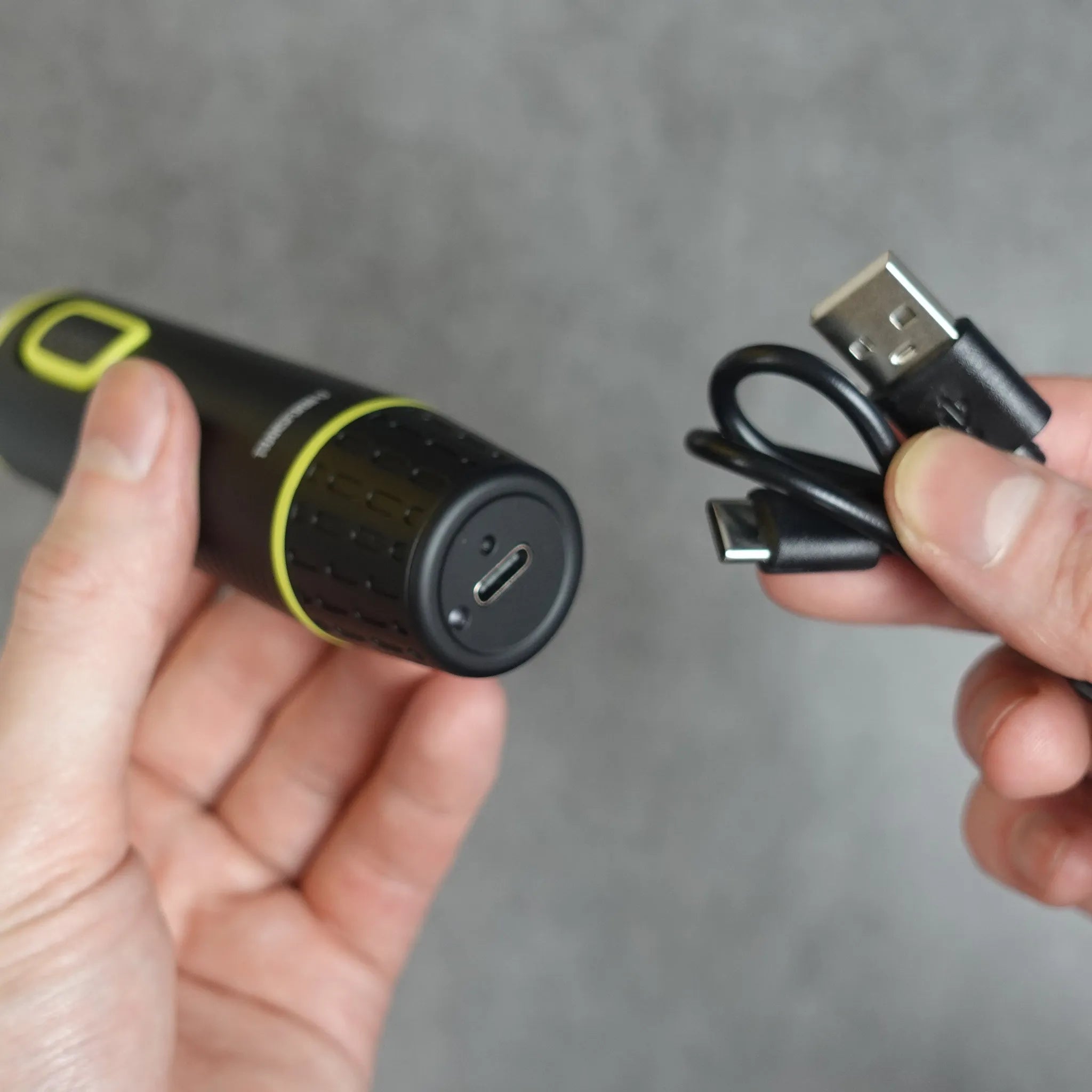Hand holding a nose and ear hair trimmer with USB cable against a gray background