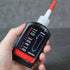 Hand holding a bottle of Blade Oil with a red cap against a gray background