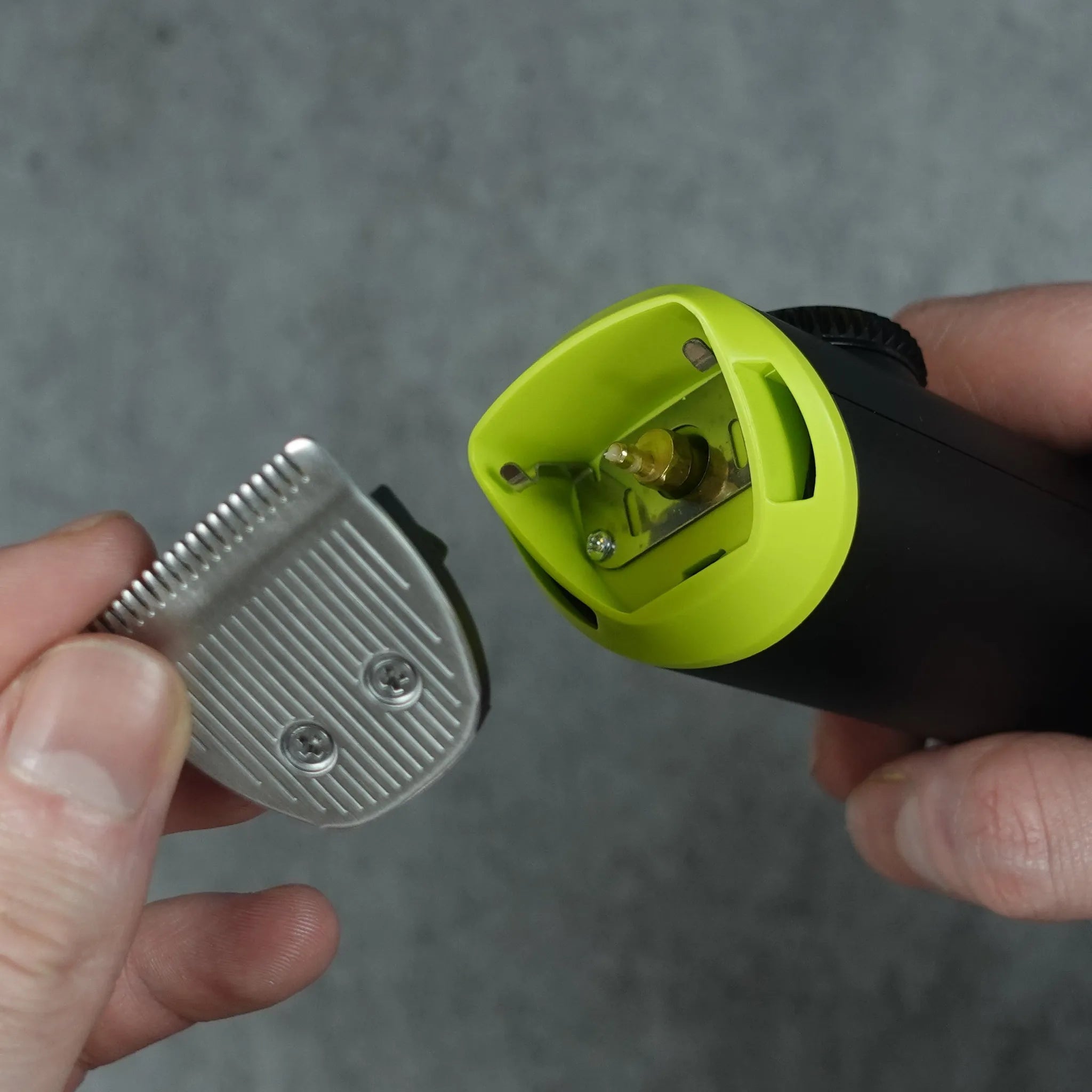 Hand holding a green and black hair trimming tool with a silver blade against a gray background