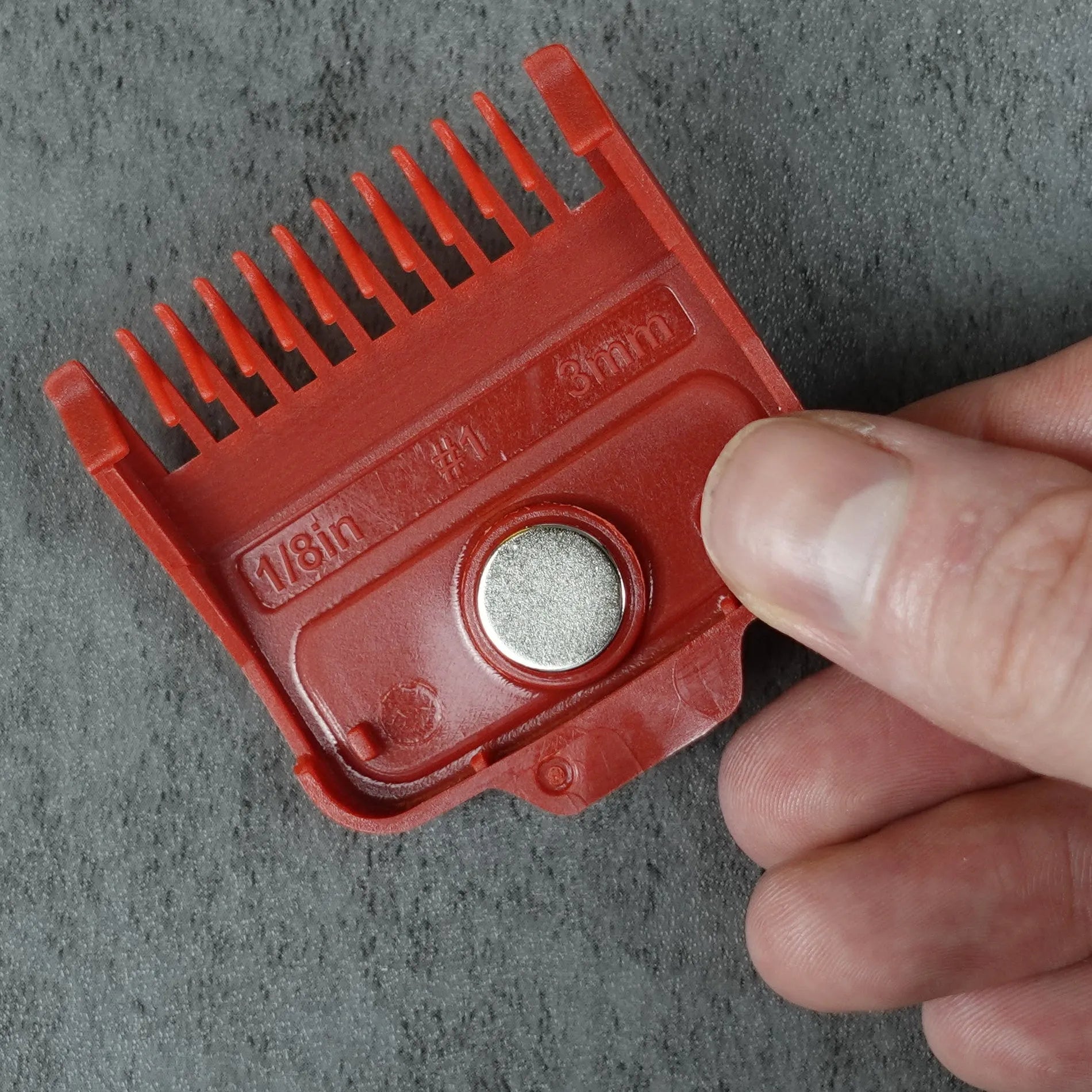 Red hair clipper magnetic guide comb held by a hand on a gray background